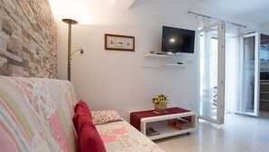 A living room with a sofa, a flat-screen TV, a small table with a vase, and a sliding glass door leading to a balcony.