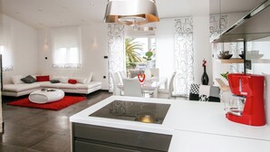 Une cuisine moderne avec un îlot central, un coin repas avec une table et des chaises blanches, et un salon avec un canapé blanc et des touches de couleur rouge.