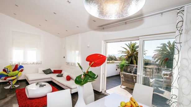 Un salon moderne avec un canapé blanc, un tapis rouge, une table blanche sur laquelle se trouve un panier de fruits, et une grande fenêtre donnant sur des palmiers et des immeubles.