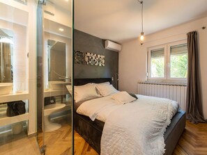 Un dormitorio moderno con cabina de ducha de vidrio, una cama grande con ropa de cama blanca, aire acondicionado de pared y una ventana con cortinas.