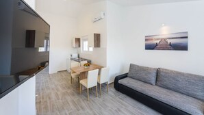 A modern living room with a grey sofa, a dining table with chairs, a kitchen area with cabinets, and a painting of a pier on the wall.