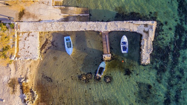 An aerial view of a coastal area with a stone pier, two boats moored at the end, and a small dock with a boat.