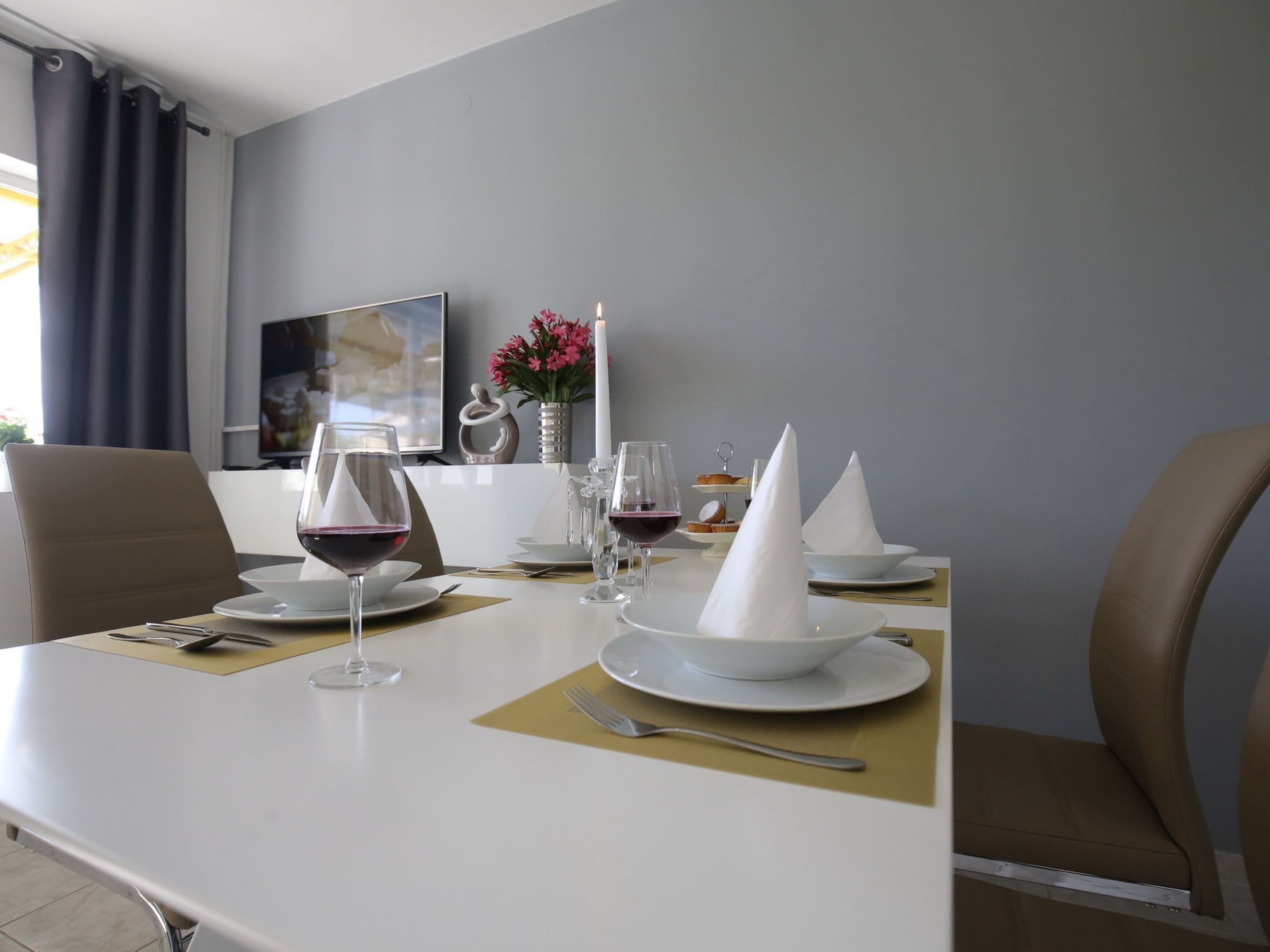 A dining table set with white plates, napkins, and wine glasses, with a vase of flowers and a candle on the table.