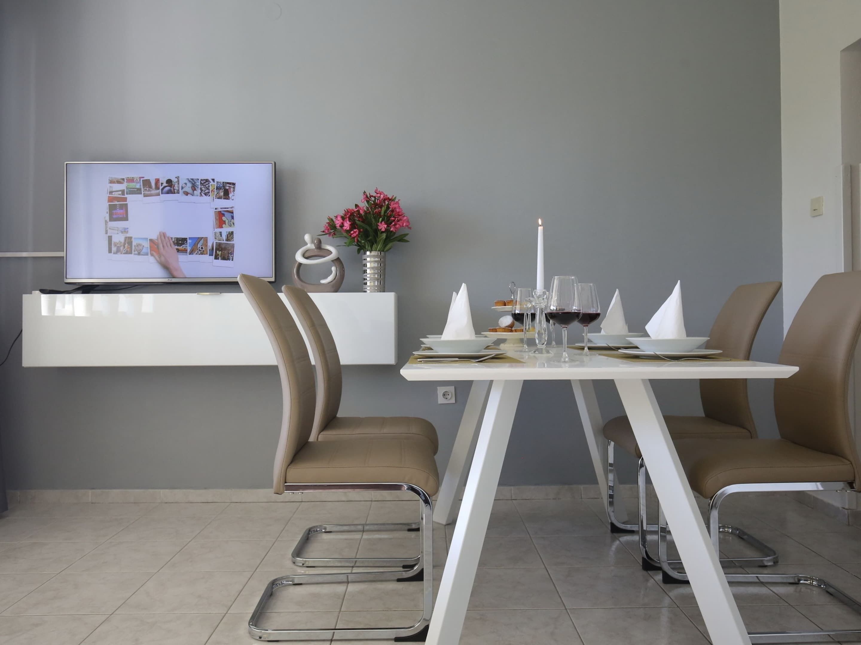 A modern dining area with a white table, two brown chairs, a TV on a white shelf, and a vase with pink flowers.