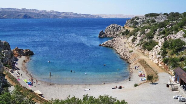 Una playa costera con aguas azules cristalinas, terreno rocoso y algunas personas disfrutando.