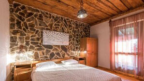 Un dormitorio con pared de piedra, techo de madera, cama con colcha blanca, armario de madera y mesita de noche con lámpara.