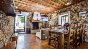 Interior rústico de piedra con una mesa y sillas de madera, una chimenea y un televisor montado en la pared.
