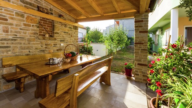 An outdoor wooden table and benches under a wooden ceiling, surrounded by stone walls and potted plants. - Luxury Apartment in Zadar With Covered Terrace (Zadar)