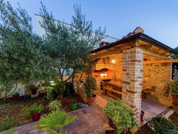 Outdoor kitchen area with stone-built oven, surrounded by lush greenery and a wooden bench.