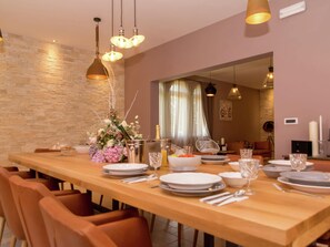 Un comedor con una larga mesa de madera dispuesta para seis personas, rodeada de sillas color naranja. Hay lámparas colgantes sobre la mesa, y una pared de piedra a la izquierda.