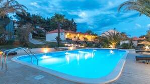 A swimming pool with a wooden deck, surrounded by palm trees and a house with a red roof.