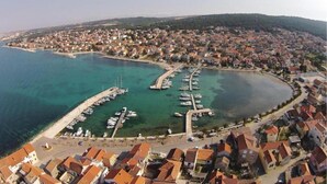 An aerial view of a coastal town with a marina, residential buildings, and a hilly landscape in the background.
