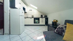 A compact kitchen with white cabinets, a microwave, and a refrigerator. There is a window providing natural light, and a sofa with a floral cushion is visible in the foreground. - Comfortable Apartment With Balcony in a Quiet Area, Near the Beach (Zadar)
