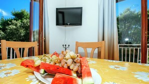 A dining table with a floral tablecloth, a plate of watermelon and grapes, and two wooden chairs. A flat-screen TV is mounted on the wall above the table.