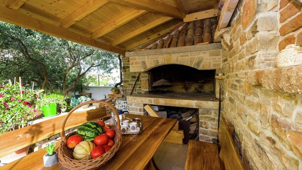 An outdoor wooden deck with a stone fireplace, a wooden table with a basket of fruit, and a bench. - New Luxury Apartment With Nice Garden and Barbecue in a Quiet Area, Wifi (Zadar)