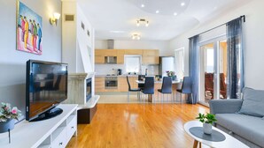 Un salon moderne doté d’un téléviseur à écran plat, d’un canapé, d’une table basse et d’un coin repas avec des chaises, et une cuisine en arrière-plan.