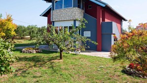 Uma casa moderna de dois andares com fachada de pedra, exterior em vermelho e azul, e um jardim bem cuidado com diversas plantas e uma pequena árvore.
