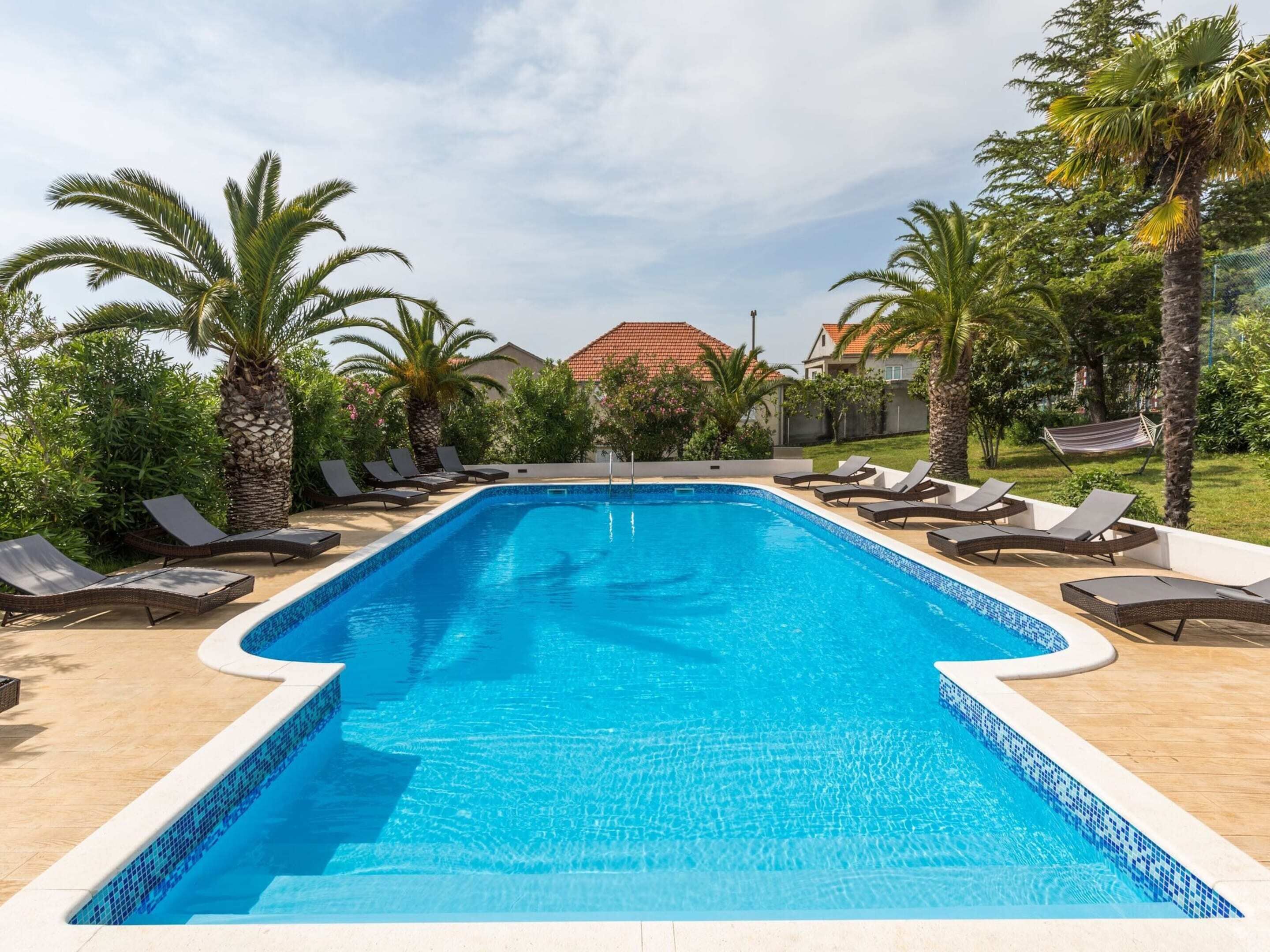 Ein rechteckiger Swimmingpool mit klarem, blauem Wasser, umgeben von Liegestühlen und Palmen.