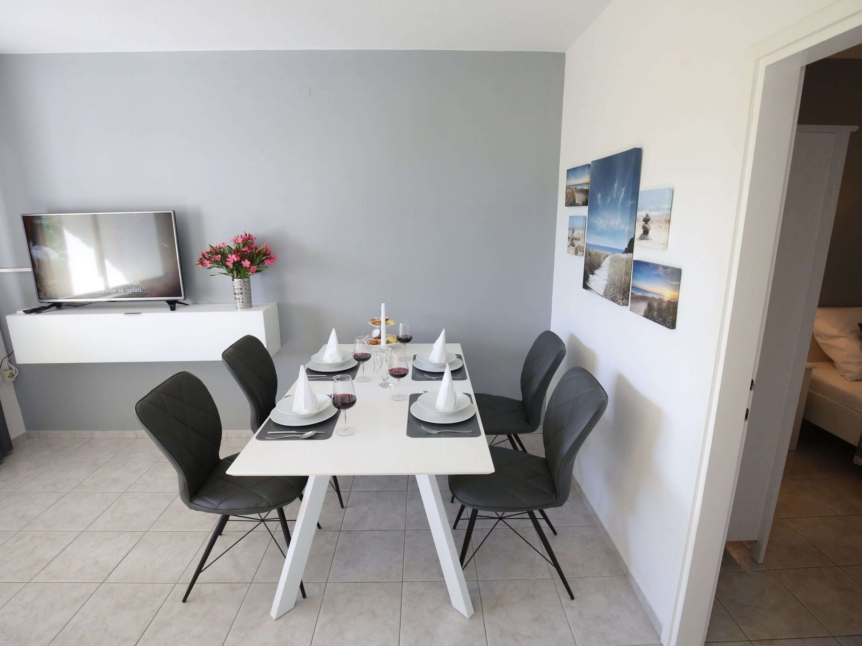A dining area with a white table set for four, black chairs, a flat-screen TV on a white stand, and a vase with pink flowers.