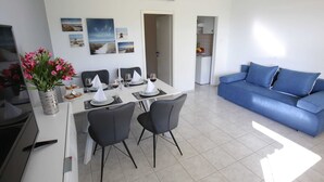 A modern living room with a blue sofa, a dining area with a white table set for four, and a kitchen visible through an open doorway. - Modern Holiday Home With Pool (Bibinje)