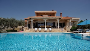 Vue sur une maison à deux étages, avec une terrasse couverte, des murs en pierre et une piscine à l’eau d’un bleu limpide, le tout vu depuis le bord de la piscine.