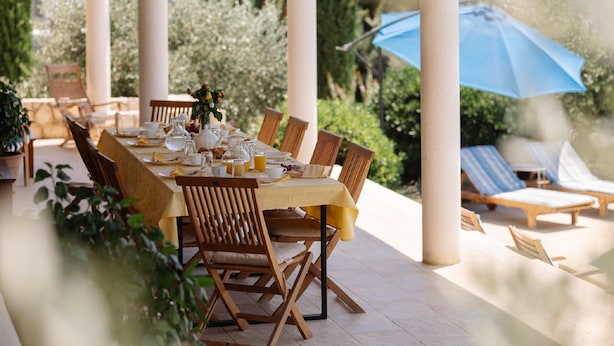 Un espace de restauration en plein air avec une table dressée pour le petit-déjeuner, entourée de chaises en bois et d’un parasol bleu.