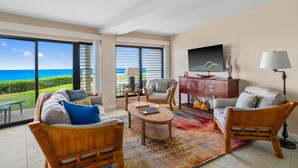 A spacious living room with a large view of the ocean, a wooden coffee table, wicker furniture, and a colorful rug.