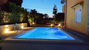 Une piscine rectangulaire avec un escalier en métal sur un de ses côtés, entourée d’un mur en pierre et éclairée par des lampes fixées au mur.
