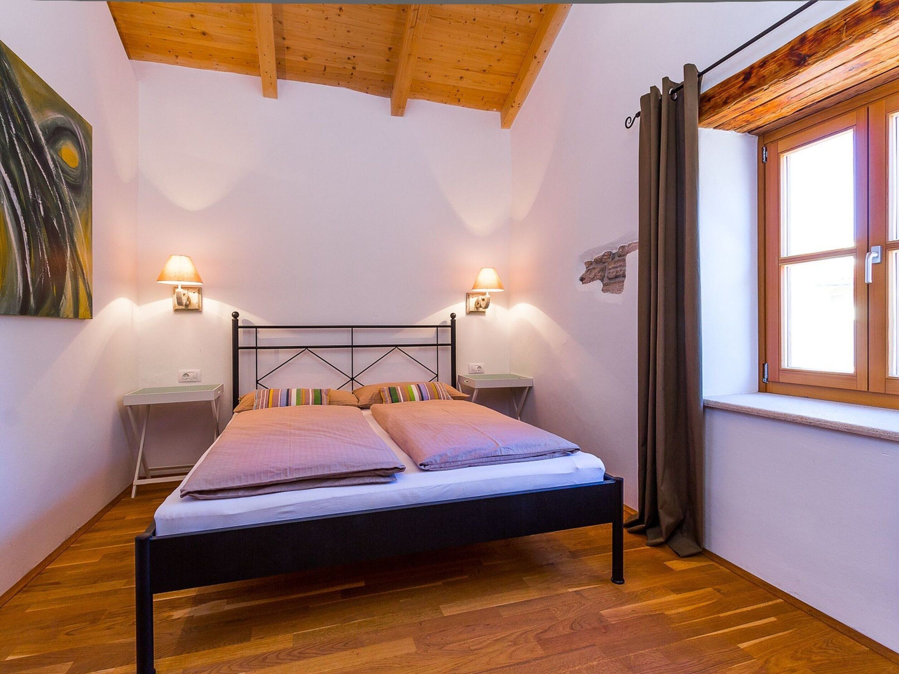 Une chambre à coucher comprenant un lit, deux tables de chevet, des lampes fixées au mur, une fenêtre avec des rideaux et un plafond en bois.