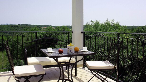 Un balcone con tavolo e sedie, un pilastro bianco e vista su vegetazione e colline.