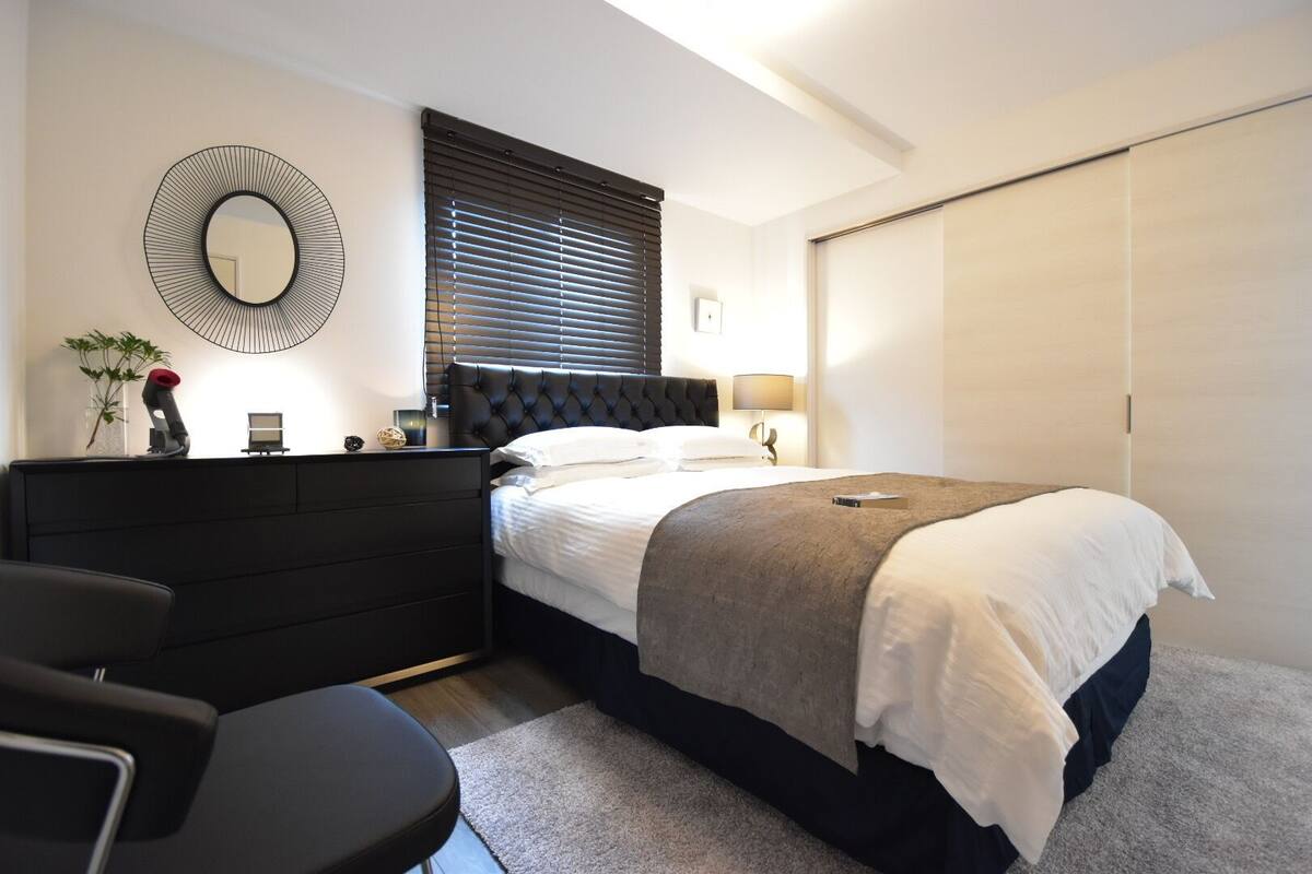 Un dormitorio moderno con una cama grande, un cabecero oscuro, un espejo redondo, una mesita de noche con una lámpara y un armario.