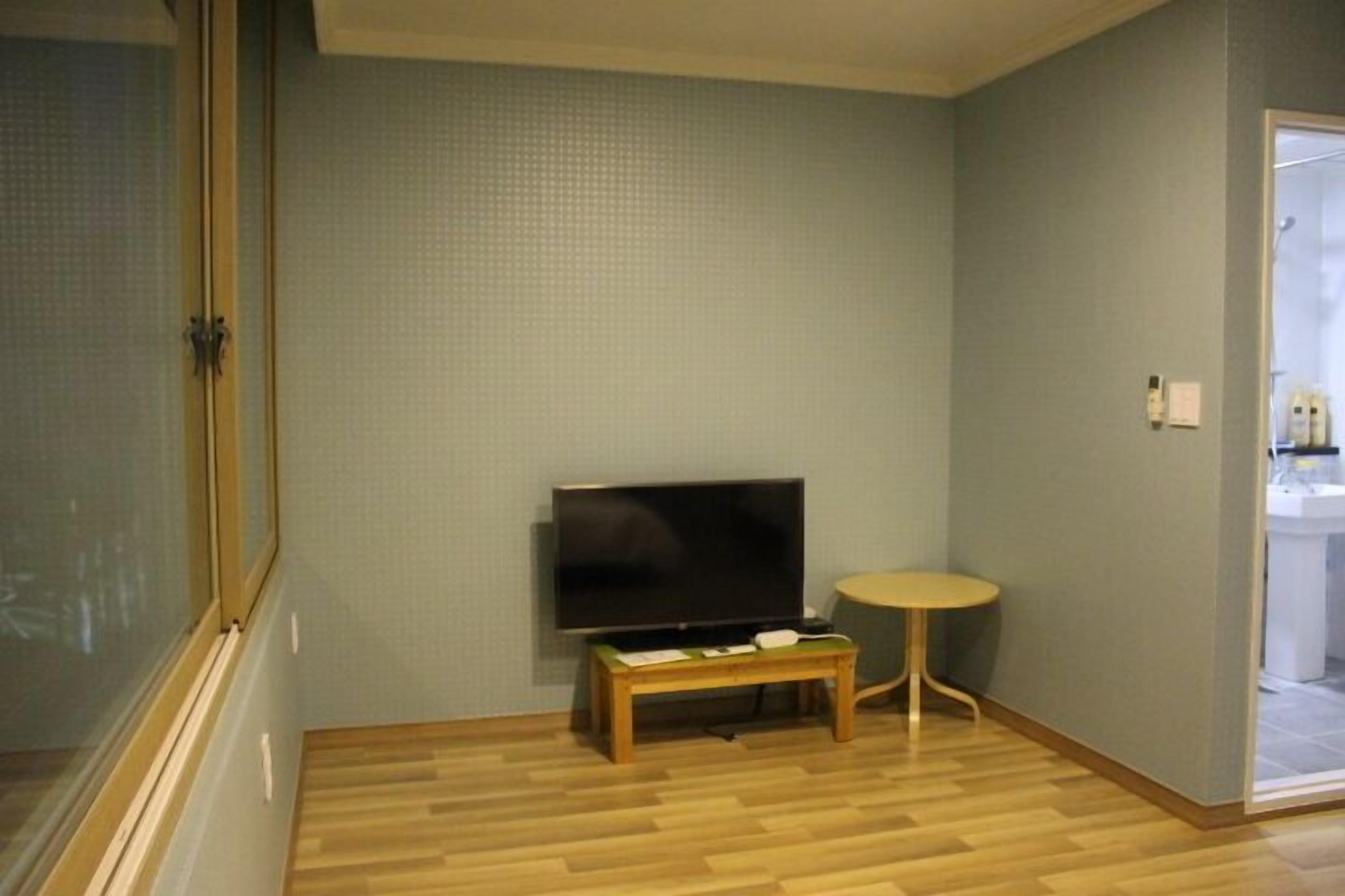 A room with a television on a wooden stand, a small round table, and a window with blinds.