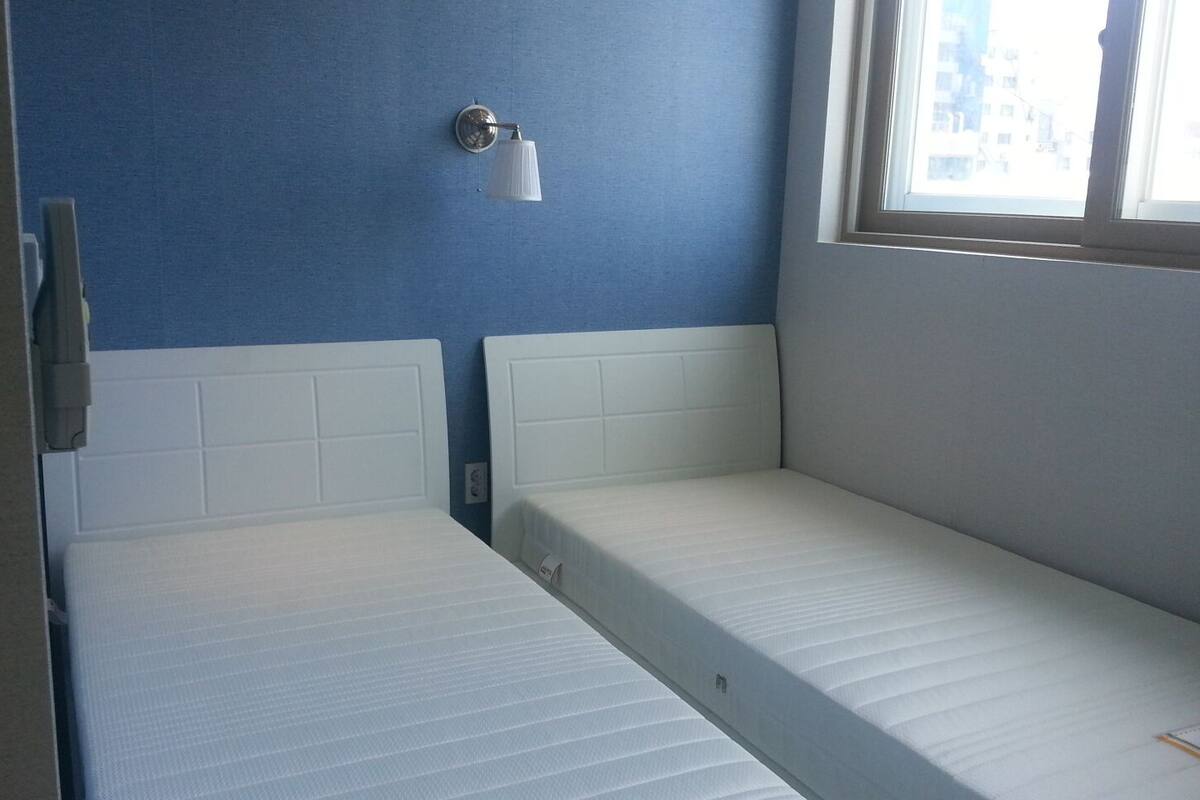 Une petite chambre avec un lit simple, une tête de lit blanche, un luminaire fixé au mur et une fenêtre donnant sur des immeubles.