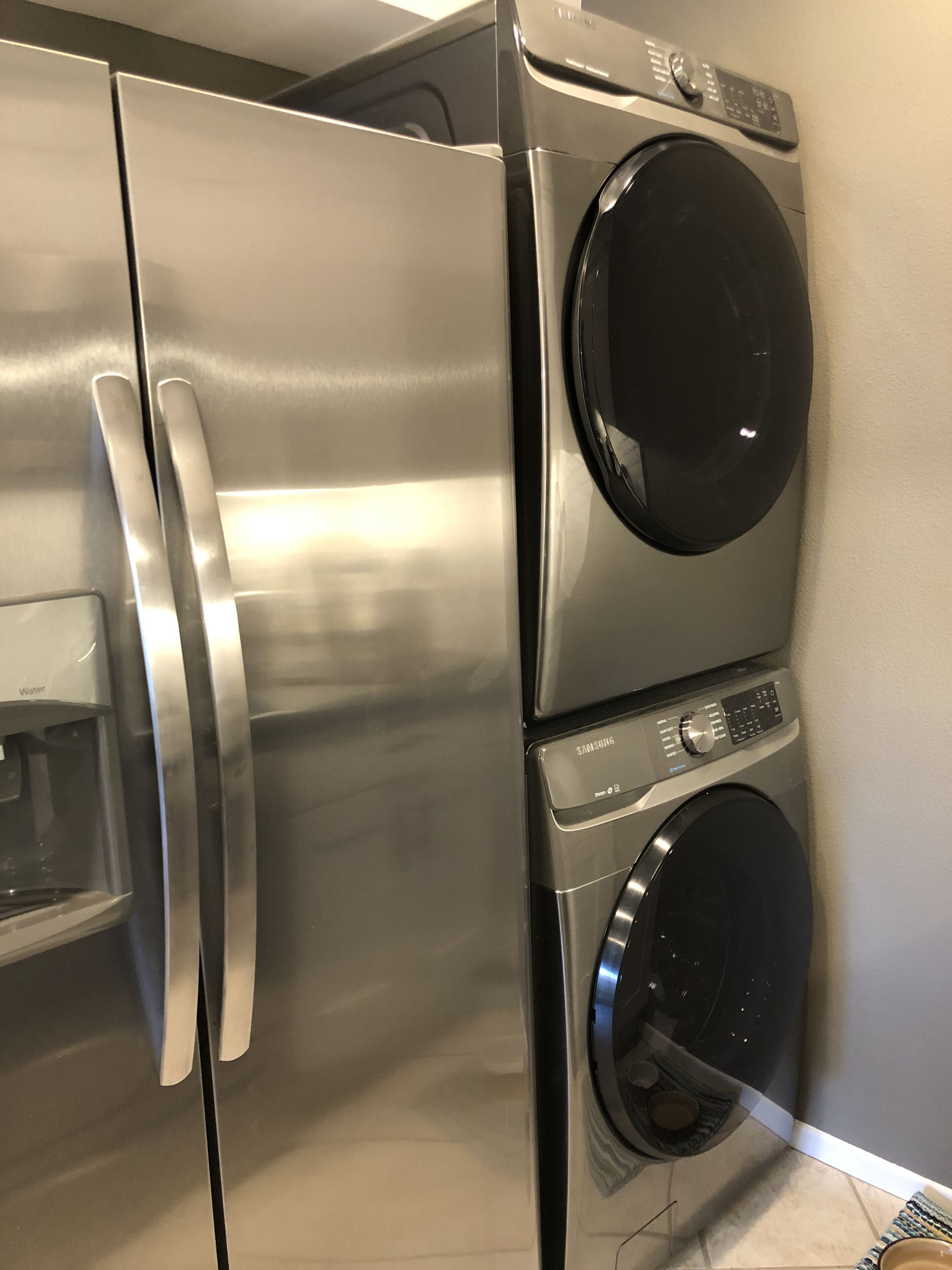 Een roestvrijstalen koelkast met een ingebouwd waterdispenser, en een verticaal geplaatst Samsung wasmachine en droger.