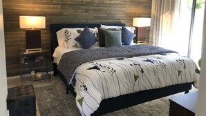 A bedroom with a wooden headboard, a bed with patterned bedding, two bedside lamps, a nightstand with decorative items, and a view of the outdoors through a window with curtains.