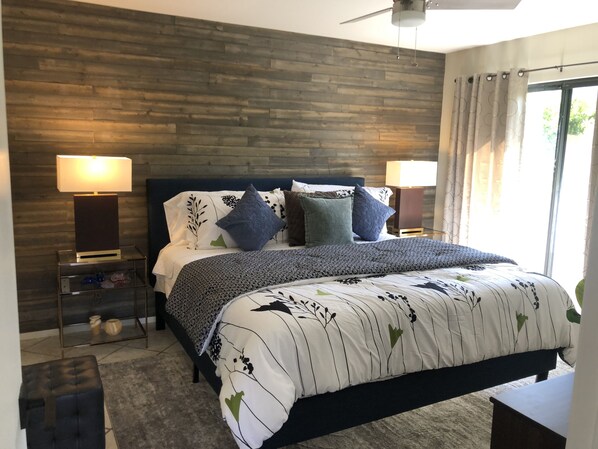 A bedroom with a wooden headboard, a bed with patterned bedding, two bedside lamps, a nightstand with decorative items, and a view of the outdoors through a window with curtains.
