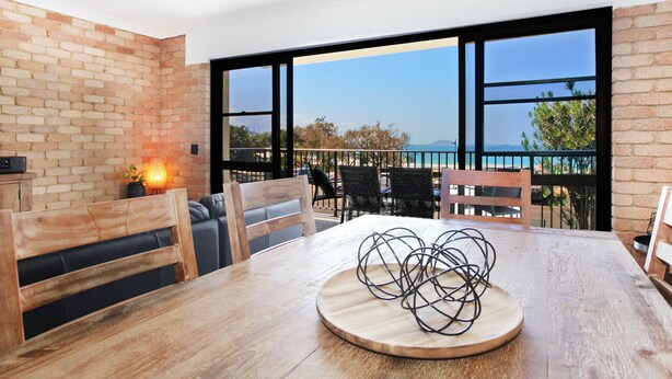A dining area with a wooden table, chairs, and a view of the sea through large windows. - Illawong 7 (Mooloolaba)