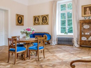 A dining room with a wooden table, blue upholstered chairs, and a vase of flowers. There are three framed portraits on the wall and a window with curtains. - Manowce Palace by Interhome (Nowe Warpno)