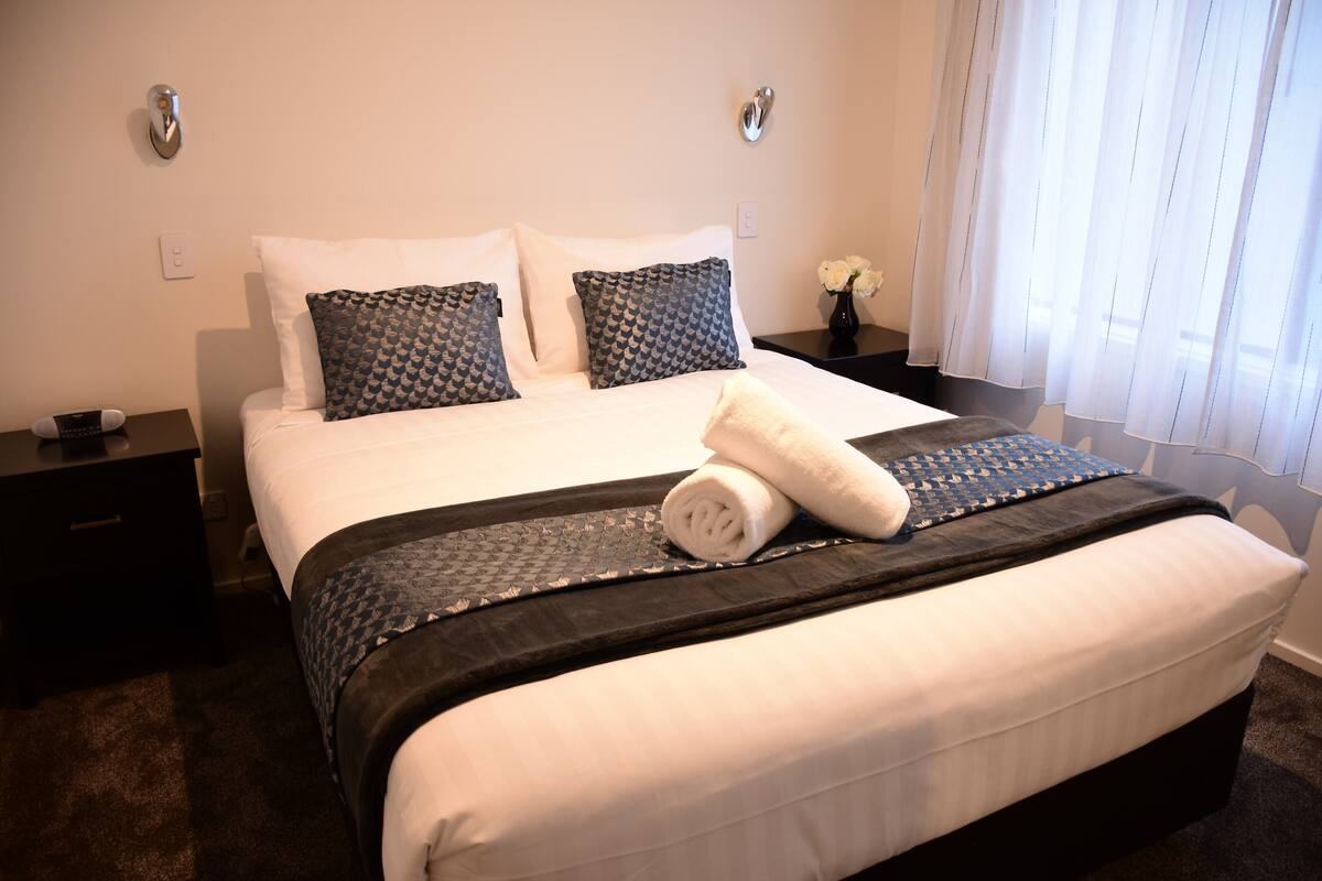 Una cama bien hecha, con almohadas blancas y estampadas, un cabecero oscuro y un corredor de cama negro.