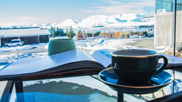 Un livre, une tasse et une table en verre, avec en toile de fond des montagnes enneigées et une voiture.