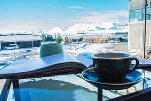 A book, a cup, and a glass table with a view of snow-covered mountains and a car in the background. - Tekapo Luxury Apartments (Lake Tekapo)