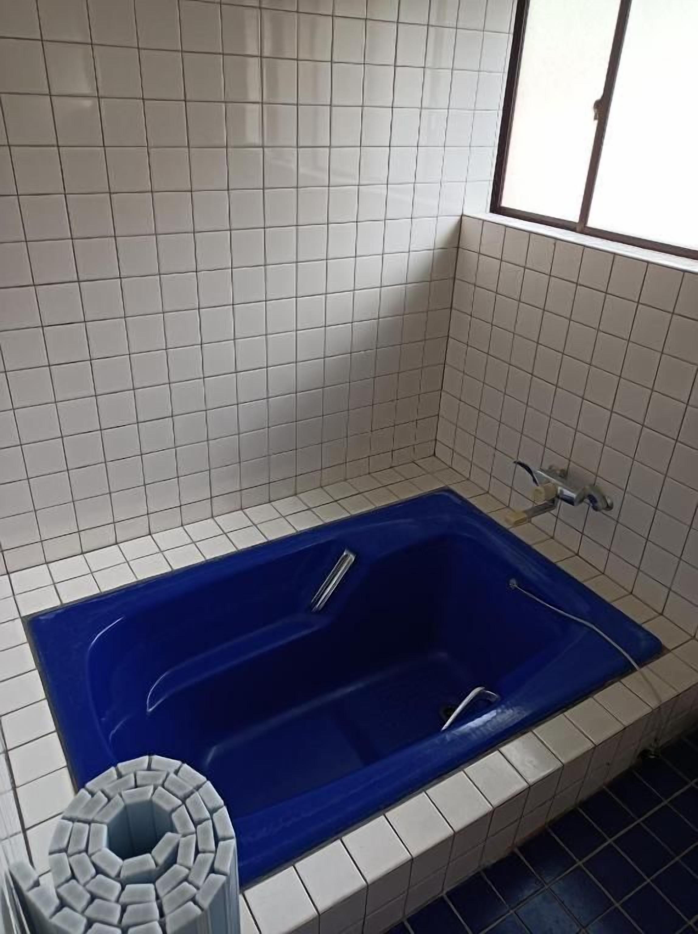 Une salle de bain avec une baignoire bleue, des murs recouverts de carreaux blancs et une fenêtre.