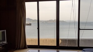 Una habitación con suelo de tatami, una mesa baja y un televisor sobre un mueble. Amplias ventanas ofrecen vistas al mar y colinas lejanas.