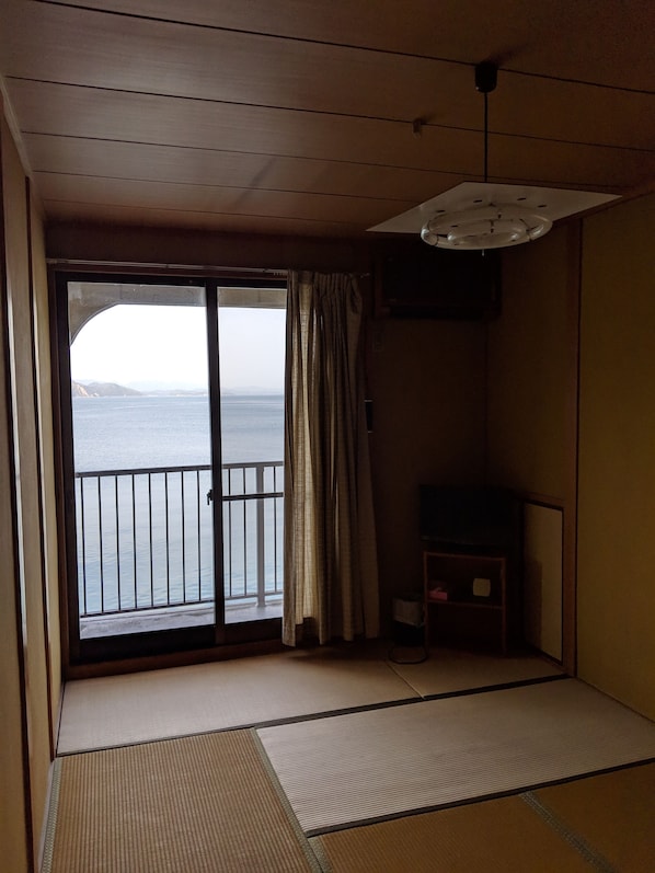 A room with tatami flooring, a sliding glass door leading to a balcony overlooking the sea, and a ceiling fan.
