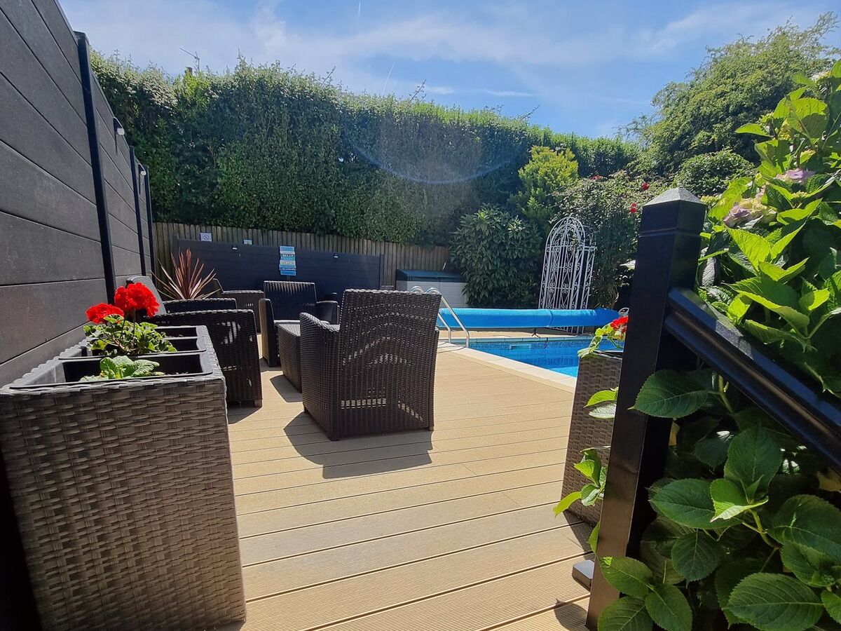 Een houten terras met rieten meubels en plantenbakken, een zwembad en een houten omheining.