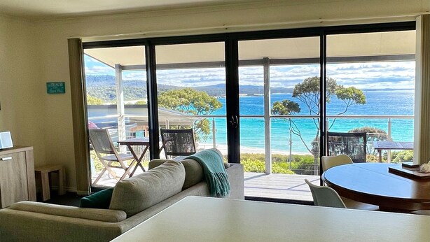 Ein Zimmer mit einer großen Schiebeglasdoor, die zu einem Balkon mit Meerblick führt. Auf dem Balkon befinden sich eine Couch, ein kleiner Tisch und ein Stuhl.