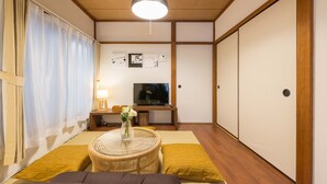 A traditional Japanese-style room with tatami flooring, a low table with a vase of flowers, a television, and sliding doors. - Yoshinaga Apartment 103 (Tokyo)