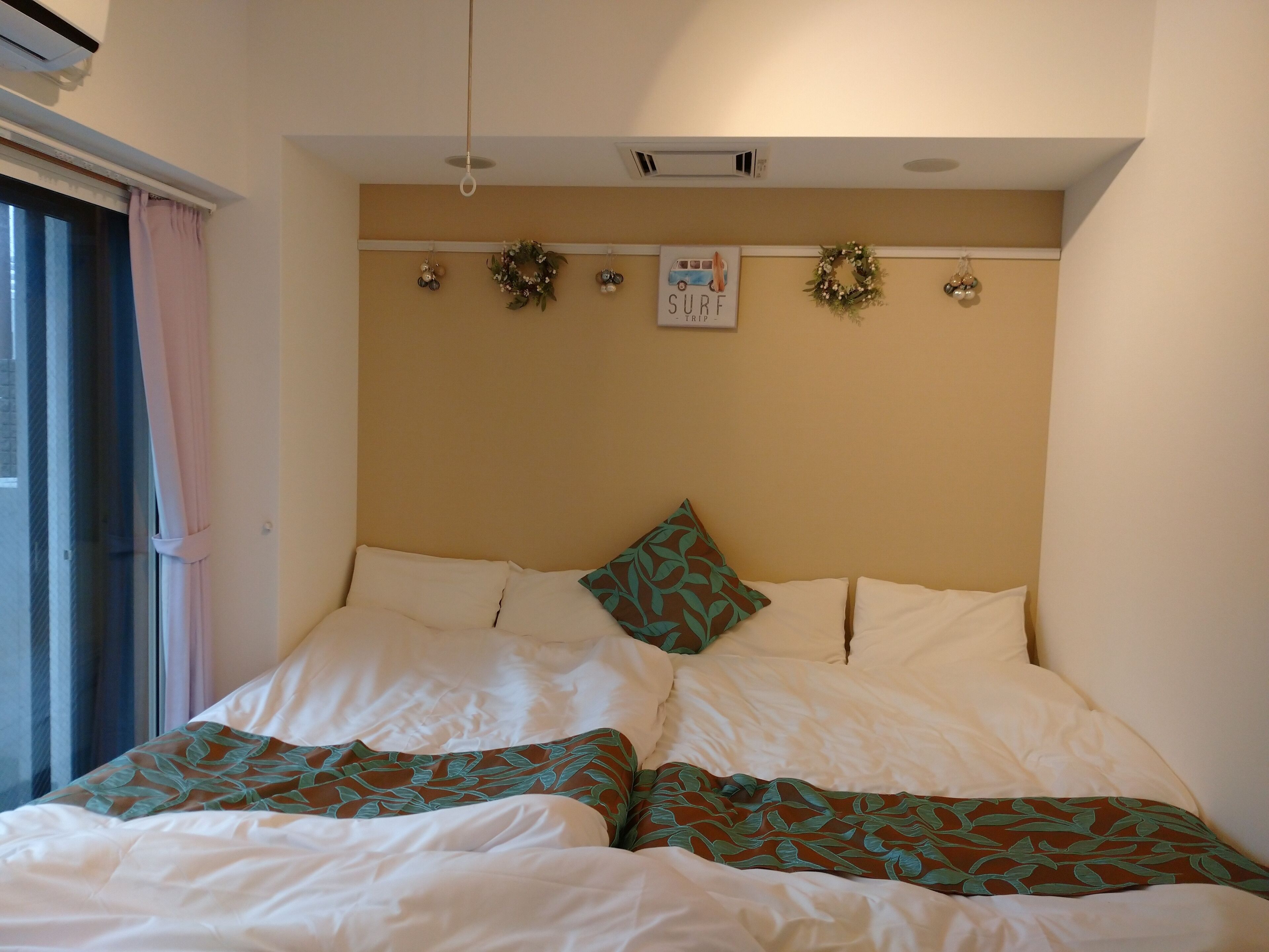 Una cama doble con sábanas blancas y cojines decorativos, un ventilador de techo y adornos en la pared, incluyendo un cartel que dice 'SURF TRIP' y guirnaldas.