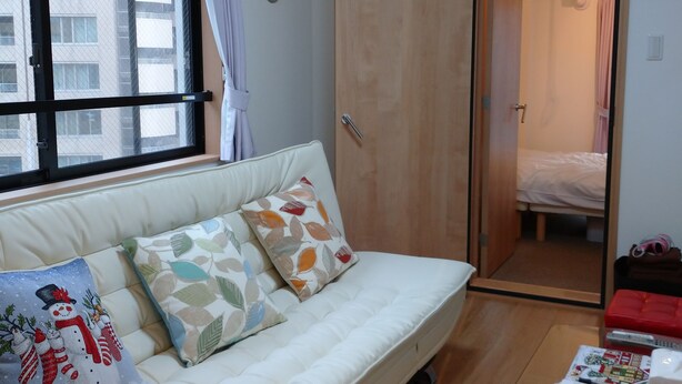 A room with a white sofa, patterned cushions, a glass table with a remote control, and a wooden door leading to a bathroom. - Lyon Doll Shinjuku Gyoen 401 (Tokyo)