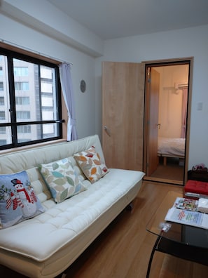 A room with a white sofa, patterned cushions, a glass table with a remote control, and a wooden door leading to a bathroom. - Lyon Doll Shinjuku Gyoen 401 (Tokyo)